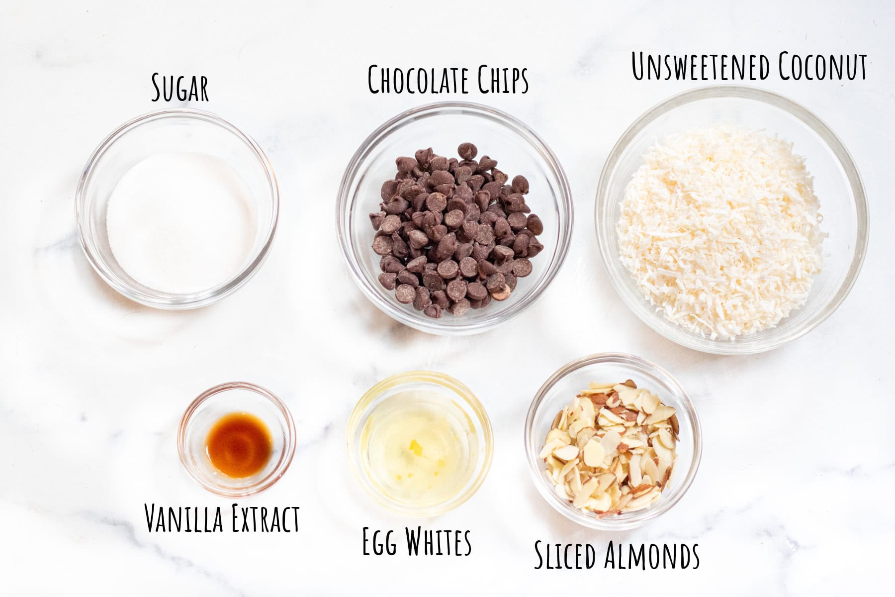 bowls of sugar, chocolate chips, coconut, vanilla extract, egg white, and sliced almonds labeled.