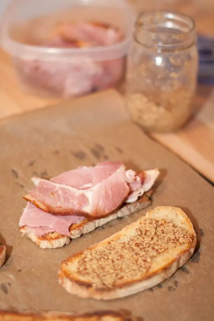 two slices of bread on parchment paper one with ham and the other with Dijon mustard.