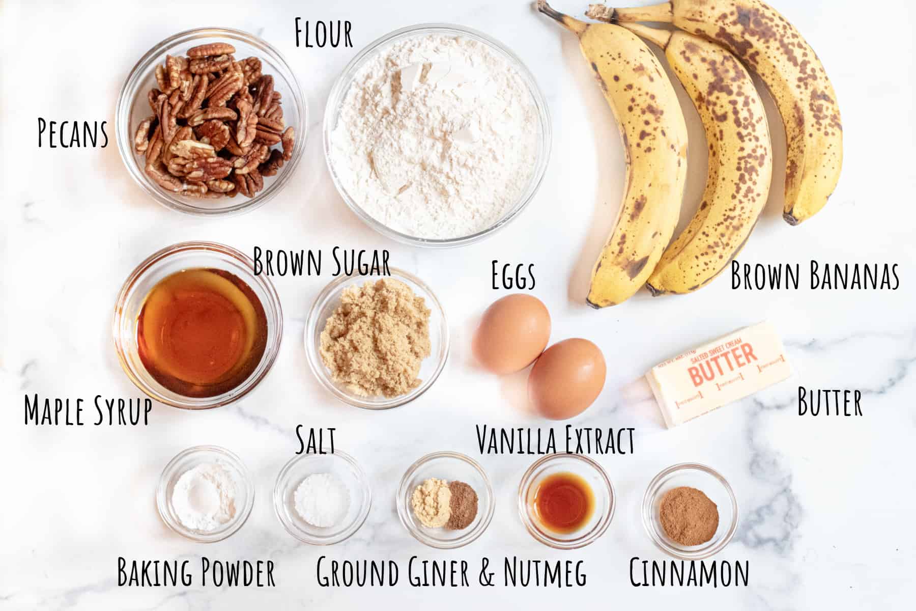 brown bananas, pecans, flour, maple syrup, brown sugar, eggs, butter, salt, baking powder, spices on a counter in bowls.