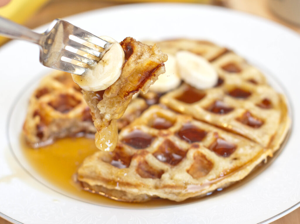 a banana waffle with syrup and a fork holding up a bite.