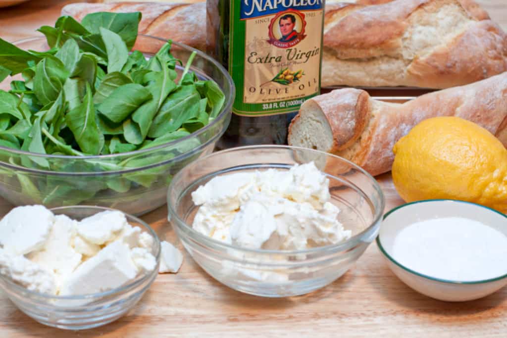 arugula, cheeses in bowls, lemon, baguette, salt, and olive oil.