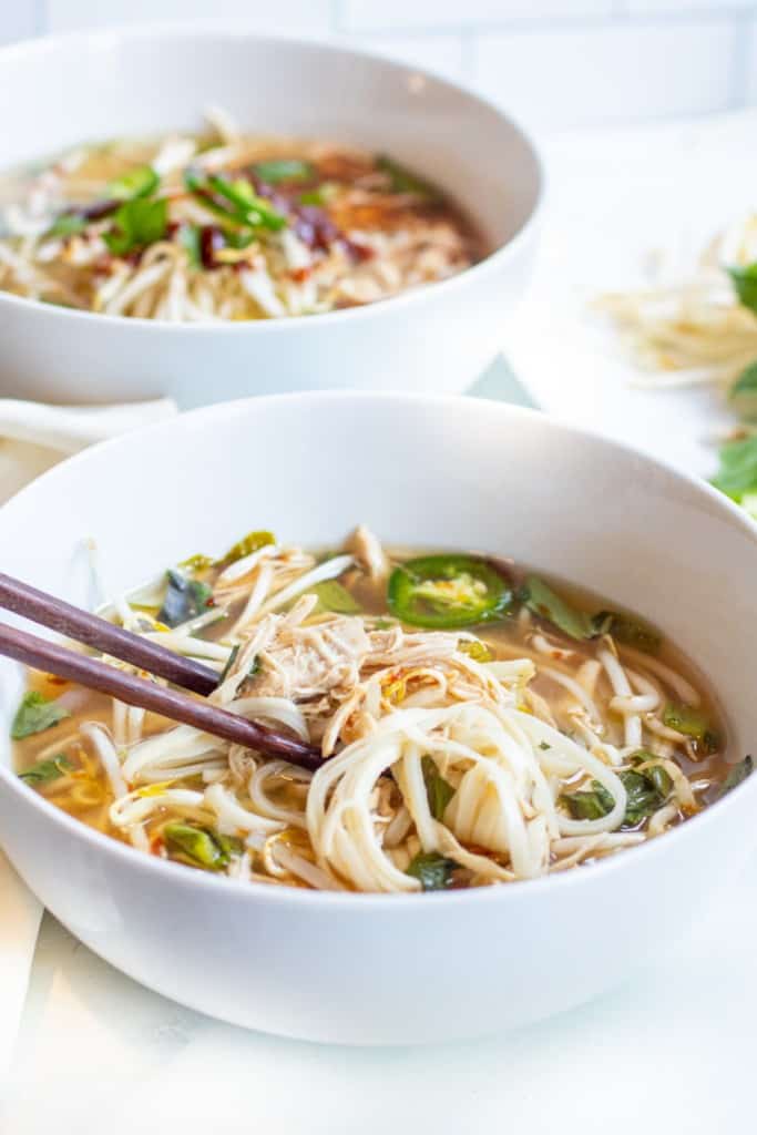 a bowl of chicken pho with chopstick holding up noodles and chicken.
