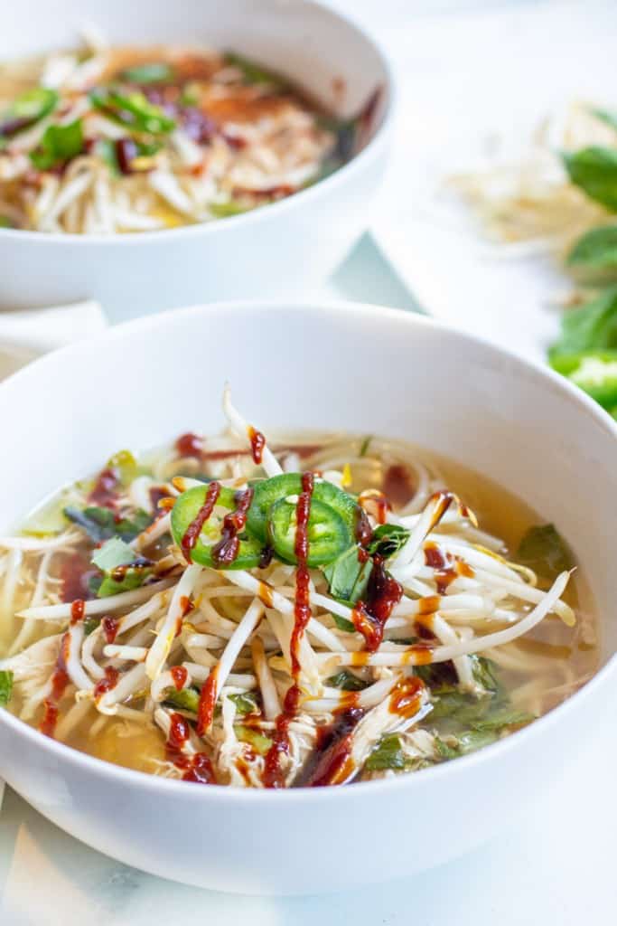 a bowl of chicken pho with sriracha, sprouts, and sliced jalapenos.