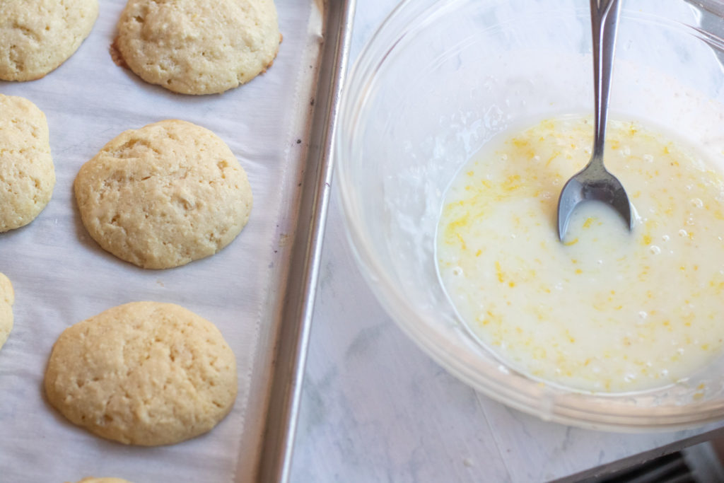 a bowl of glaze with spoon next to baked cookies.