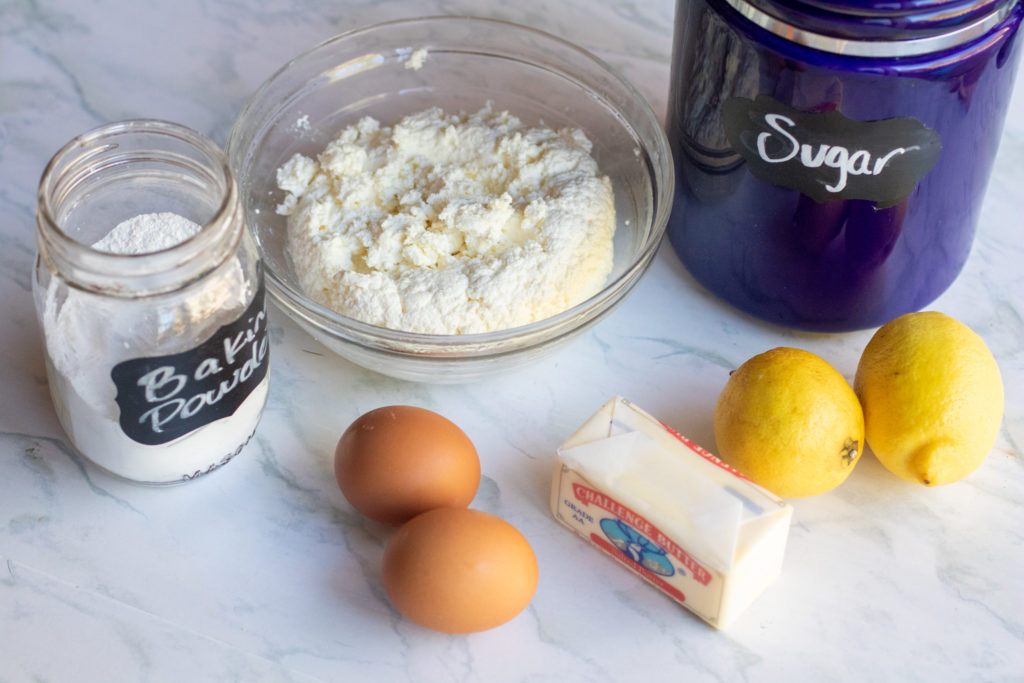 cheese, baking powder, sugar, butter, lemons, eggs on a counter.