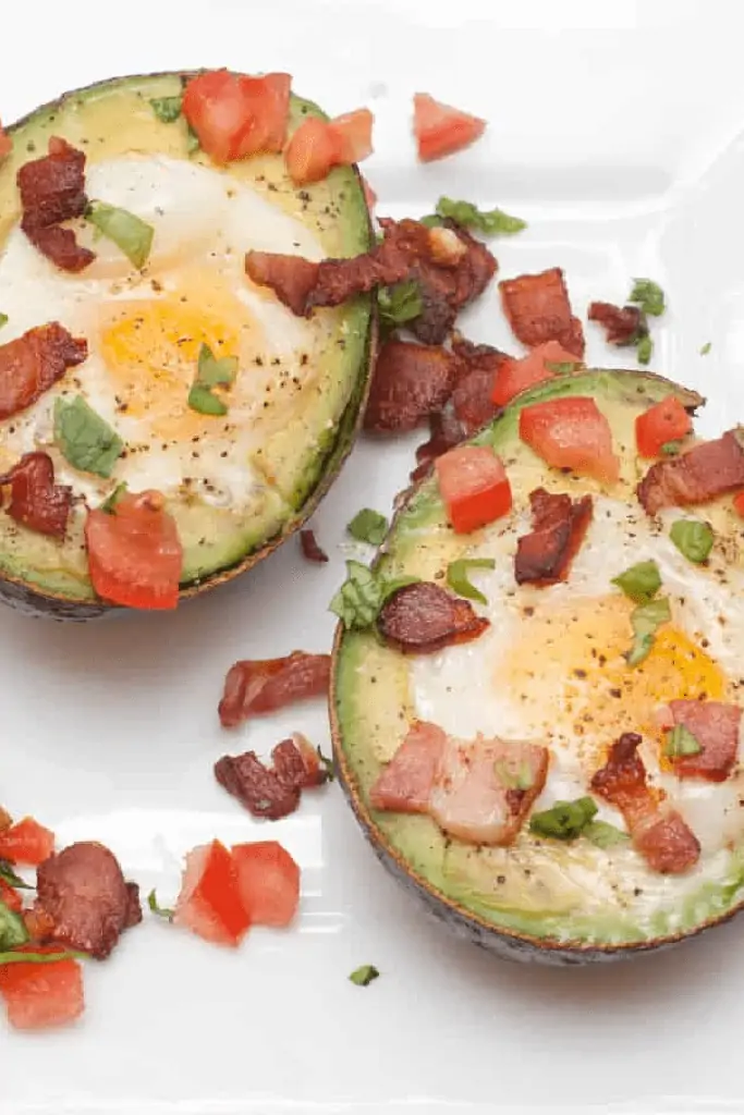 a plate with baked eggs in avocado topped with crumbled bacon and tomatoes.