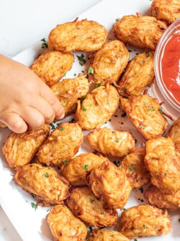 a plate of homemade tater tots with ketchup and a little hand grabbing one.