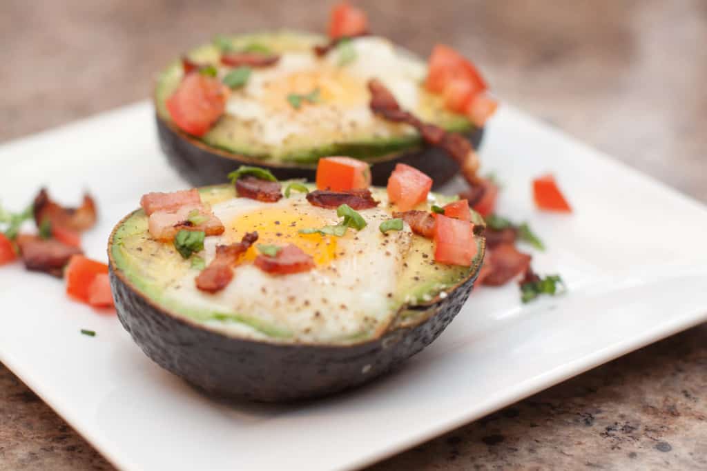 a plate with halved avocado eggs topped with baked eggs and bacon and tomatoes.