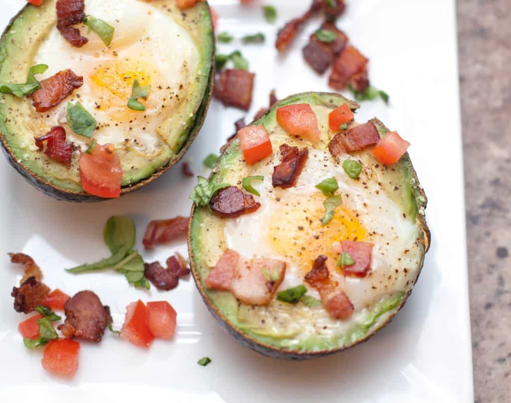 a plate with halved avocado eggs topped with baked eggs and bacon and tomatoes.