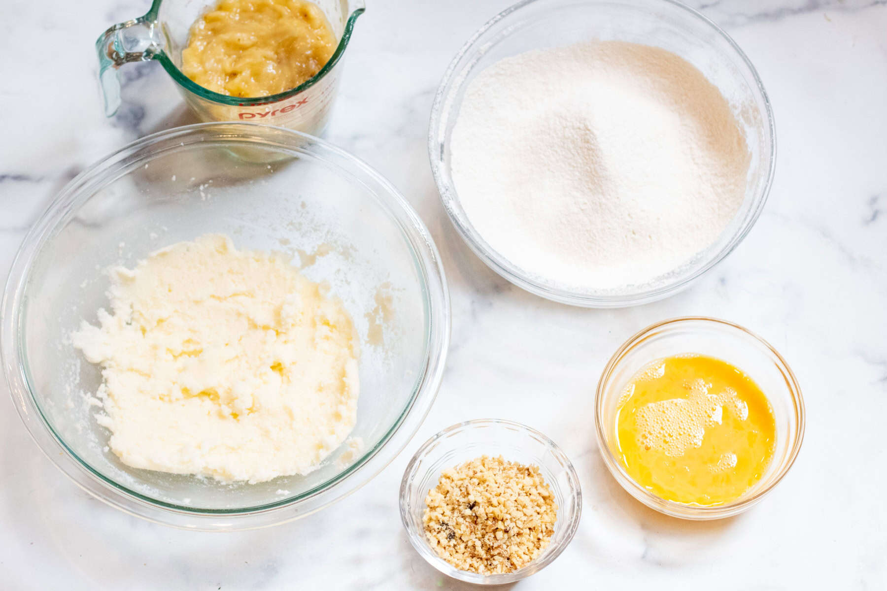 mashed bananas in a bowl, sifted flour, creamed sugar and butter, walnuts, and whisked eggs in a bowl.