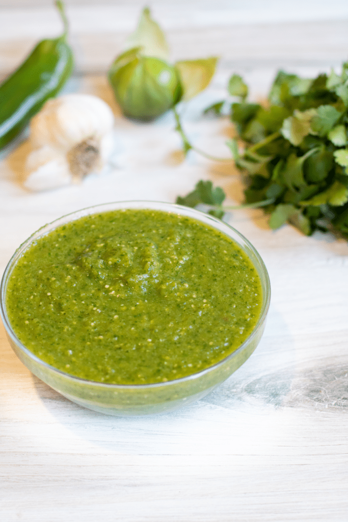 a bowl of green salsa with peppers and cilantro behind it.