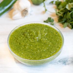 a bowl of green salsa with peppers and cilantro behind it.