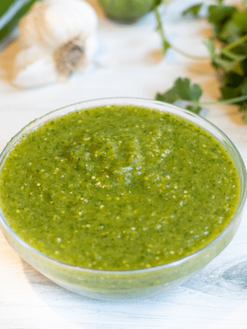 a bowl of green salsa with peppers and cilantro behind it.