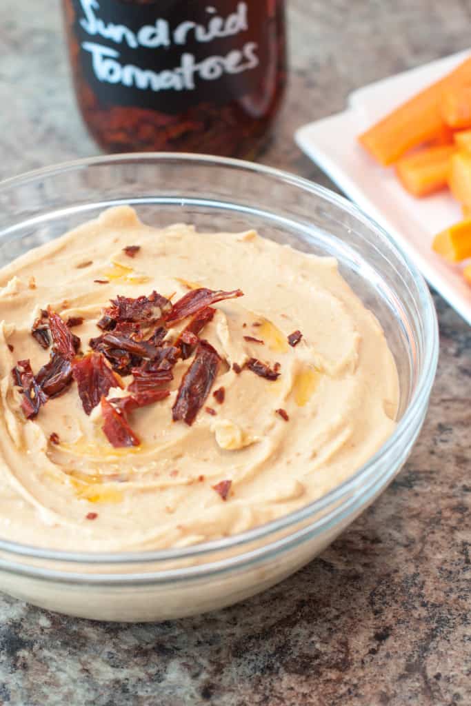 a bowl of hummus with sun-dried tomatoes in front of carrots.