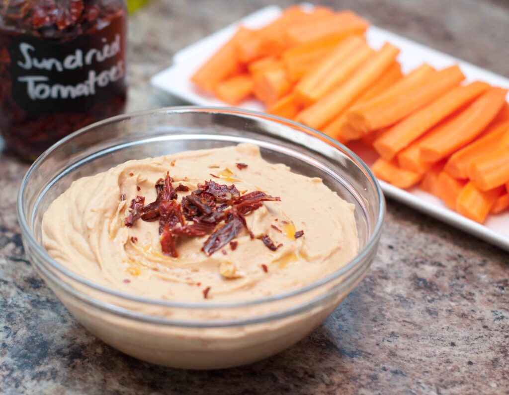 a bowl of hummus with sun-dried tomatoes in front of carrots.