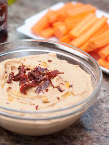 a bowl of hummus with sun-dried tomatoes in front of carrots.