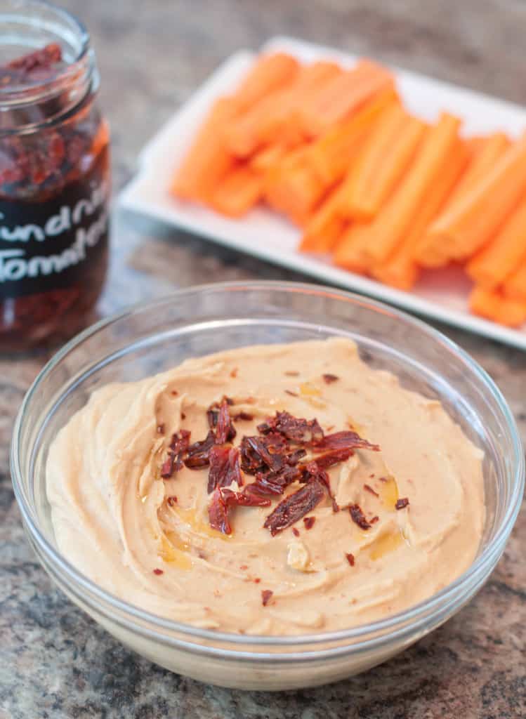 a bowl of hummus with sun-dried tomatoes in front of carrots.