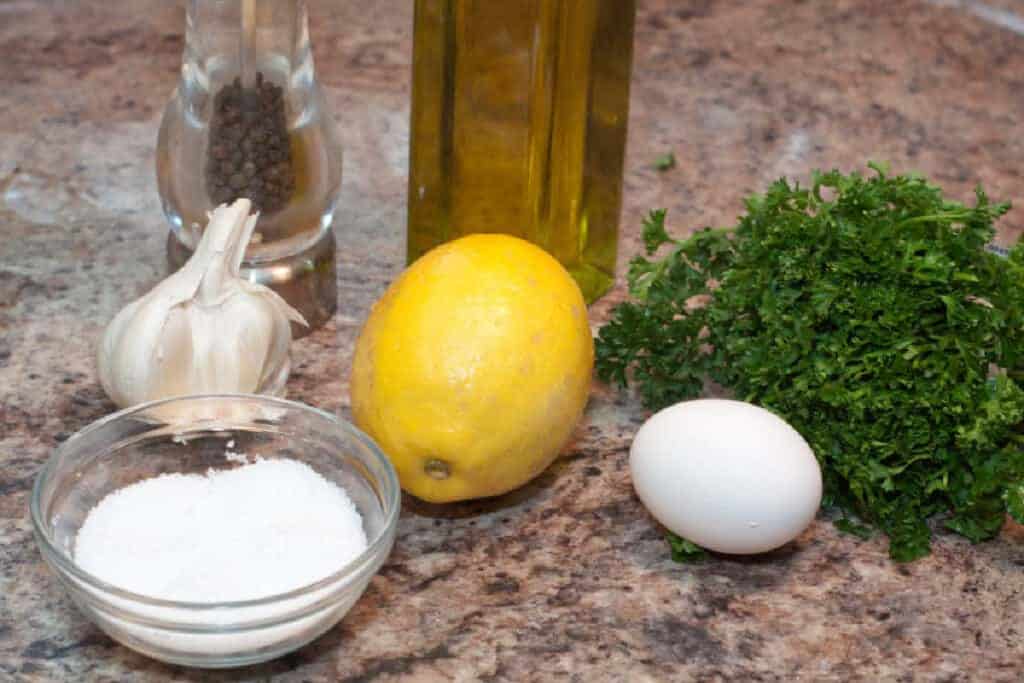 lemon, egg, parsley, oil, salt, garlic, pepper on a counter.