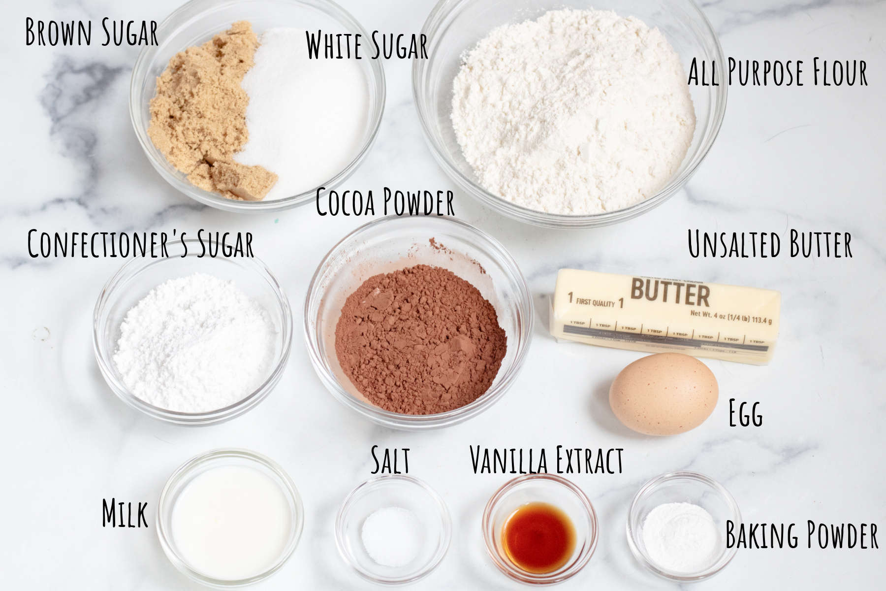 a bowl of brown and white sugar, flour, cocoa, butter, egg, confectioner's sugar, vanilla extract, milk, and baking powder and salt on a counter.