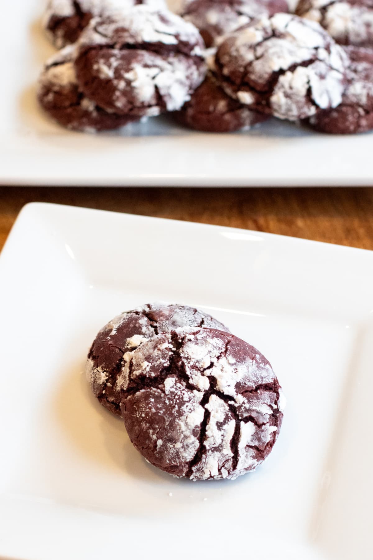 two red velvet cookies on a plate in front of a larger plate of cookies. 