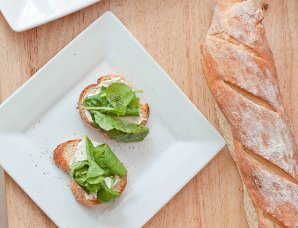 a plate with sliced baguettes topped with cheese and arugula next to a baguette.