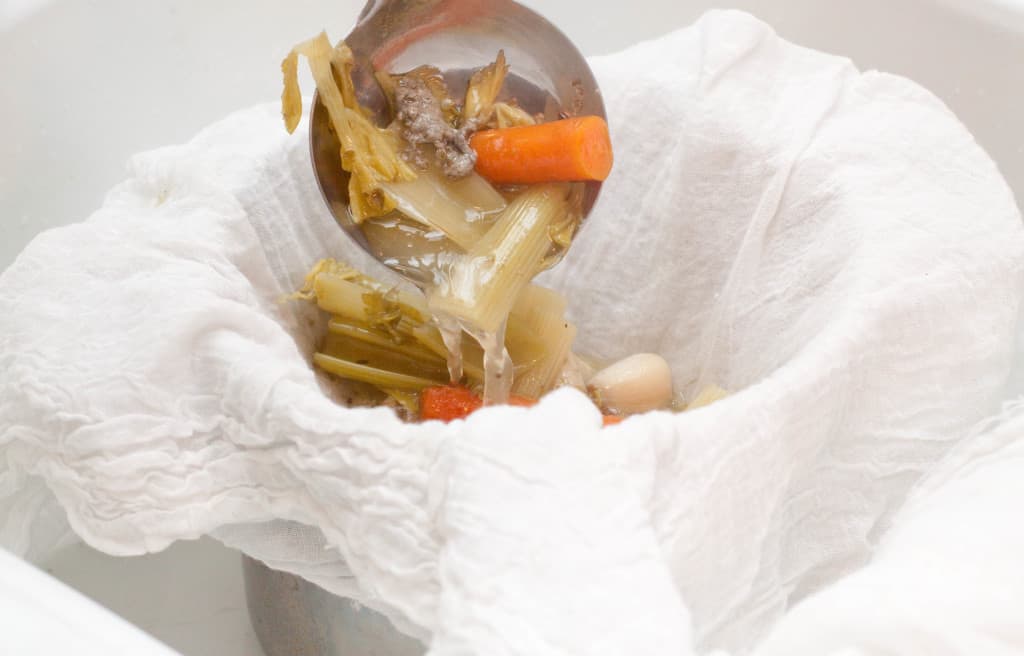 a ladle pouring celery, carrots, and stock into a cheese cloth.