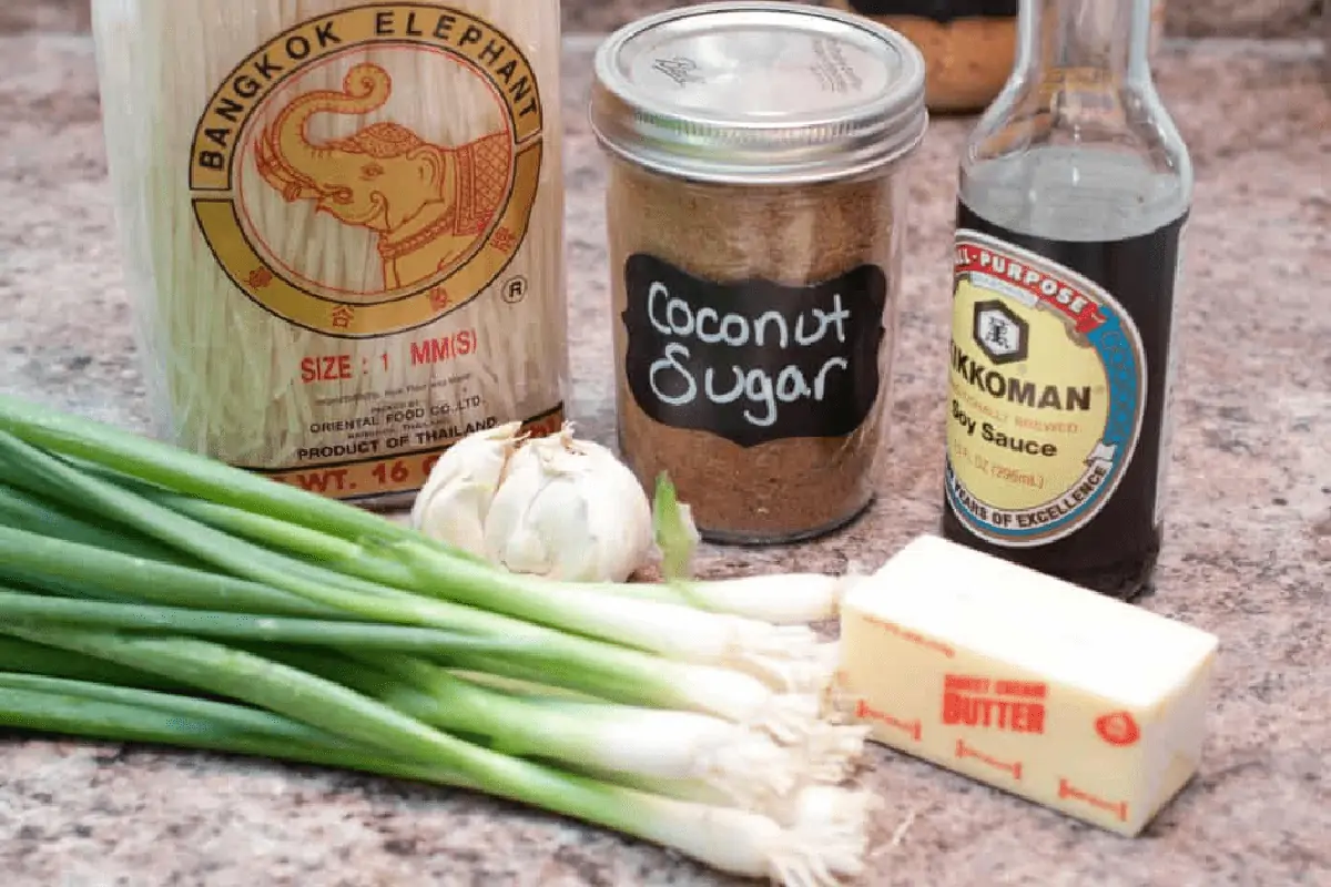 rice noodles, green onions, coconut sugar, soy, butter, garlic on a counter.