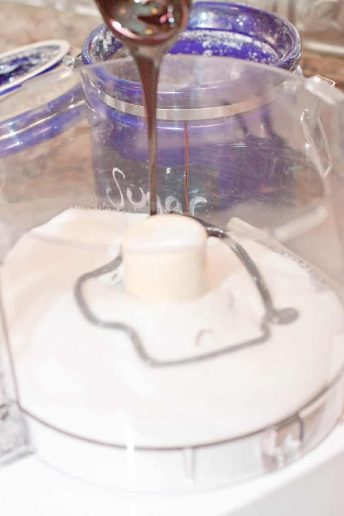 molasses being poured into a food processor bowl with white sugar.