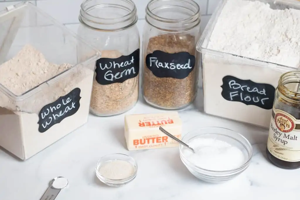 flours, jars of wheat germ and flax seed, sugar, butter, salt, and malt barley syrup on counter.