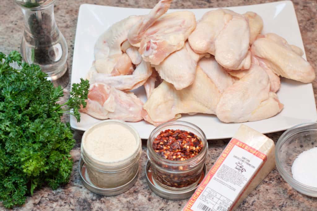 a plate of raw chicken wings, parsley, red pepper flakes, cheese garlic powder salt and pepper on a counter