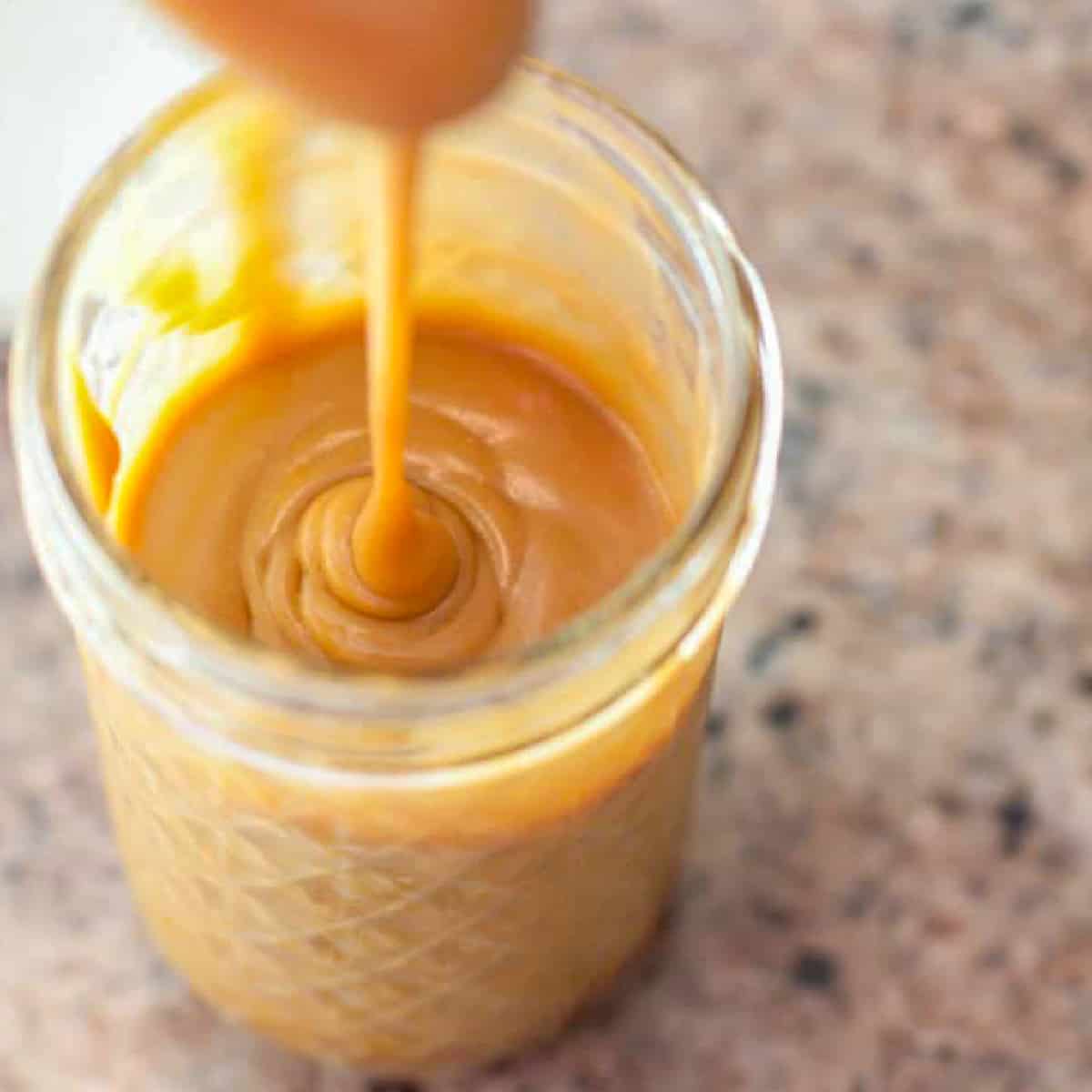 an open mason jar of caramel sauce and a spoon pouring it in.