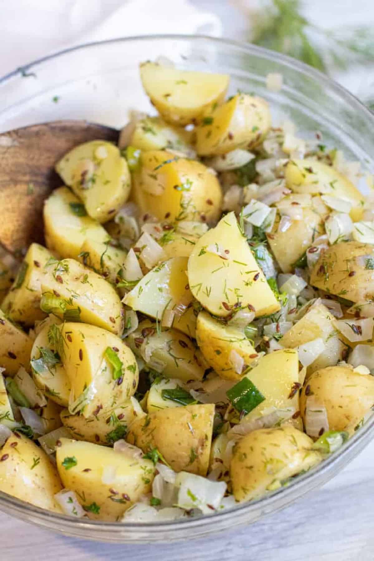 a bowl of yellow potato salad with seeds, onions, and dill with a wooden spoon.
