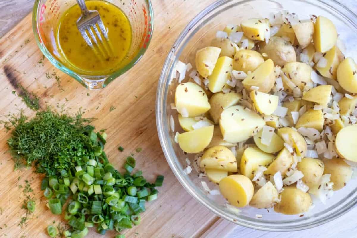 a cutting board with diced green onions, dill, a vinaigrette, and a bowl of potato salad with onions.