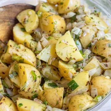 a bowl of yellow potato salad with seeds, onions, and dill with a wooden spoon.