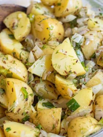 a bowl of yellow potato salad with seeds, onions, and dill with a wooden spoon.