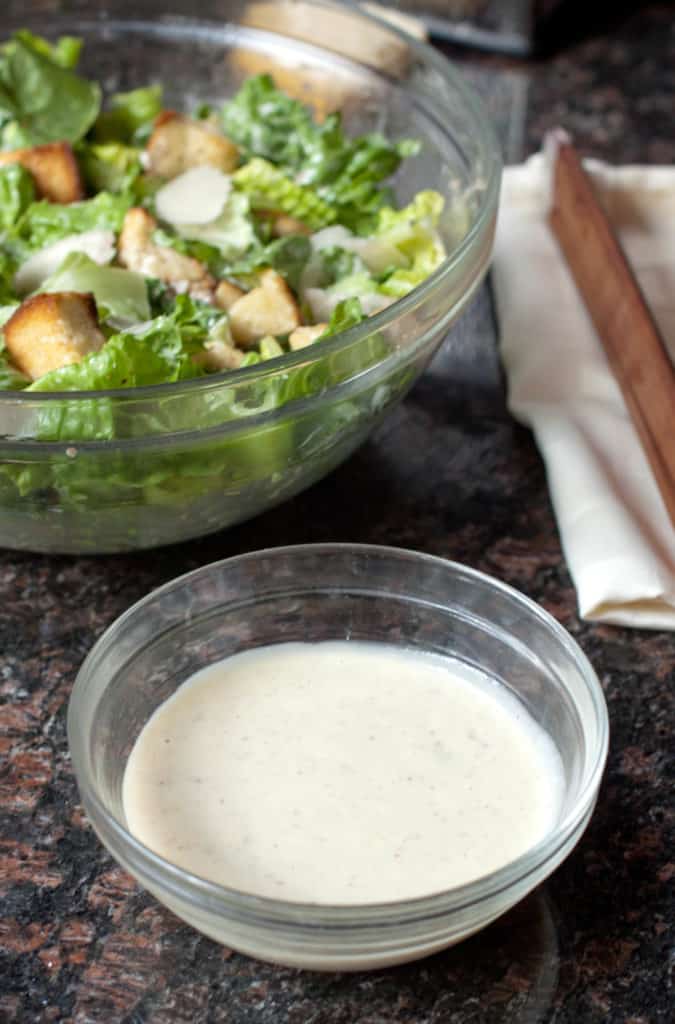 a small bowl of dressing in front of a big bowl of caesar salad.