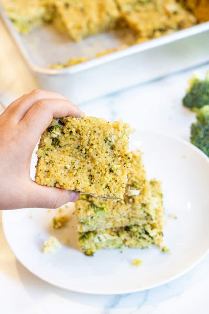 a kid's hand holding a quinoa broccoli cheddar bar over a plate of them.