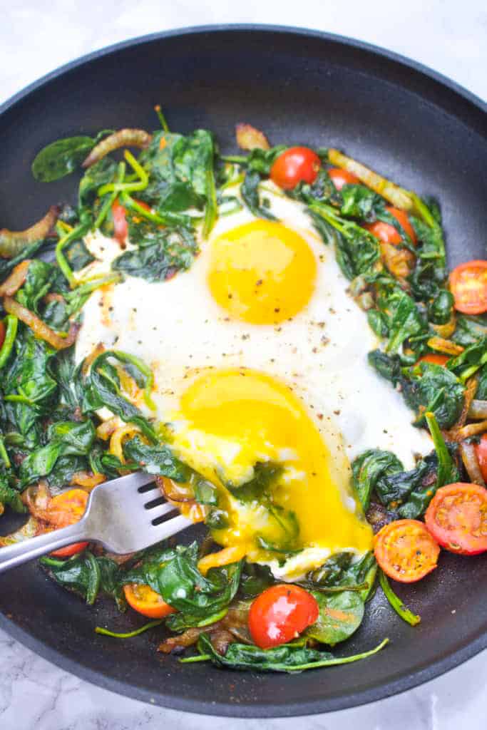 a pan with sauteed spinach, tomatoes, onions, and eggs with runny yolk.