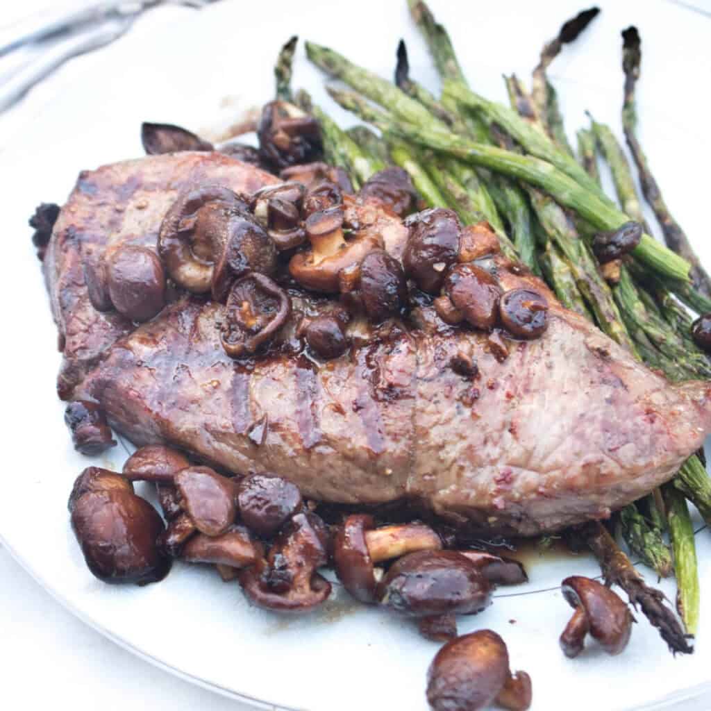 a plate of grilled steak topped with cooked mushrooms with asparagus on the side.