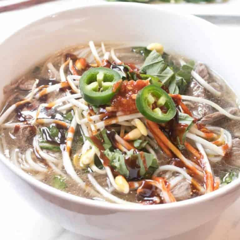a bowl of beef pho with bean sprouts, jalapenos, basil, and sriracha.