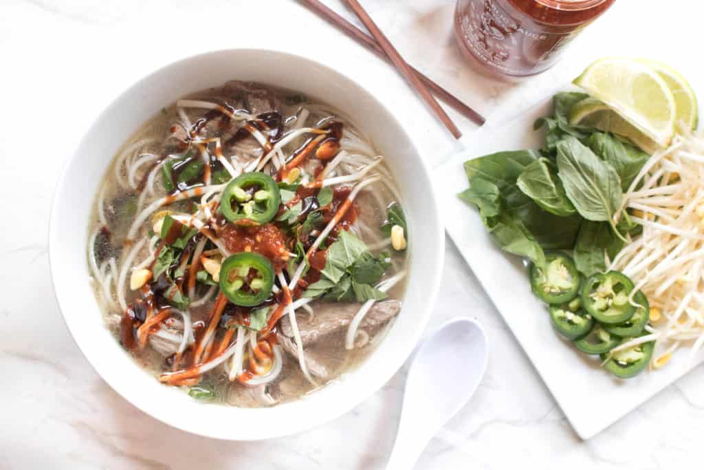 overhead of a bowl of pho and a plate of basil, jalapenos, and bean shoots next to it.