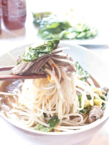 a bowl of beef pho with chopsticks holding a bite up