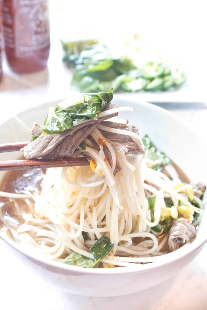 a bowl of beef pho with chopsticks holding a bite up