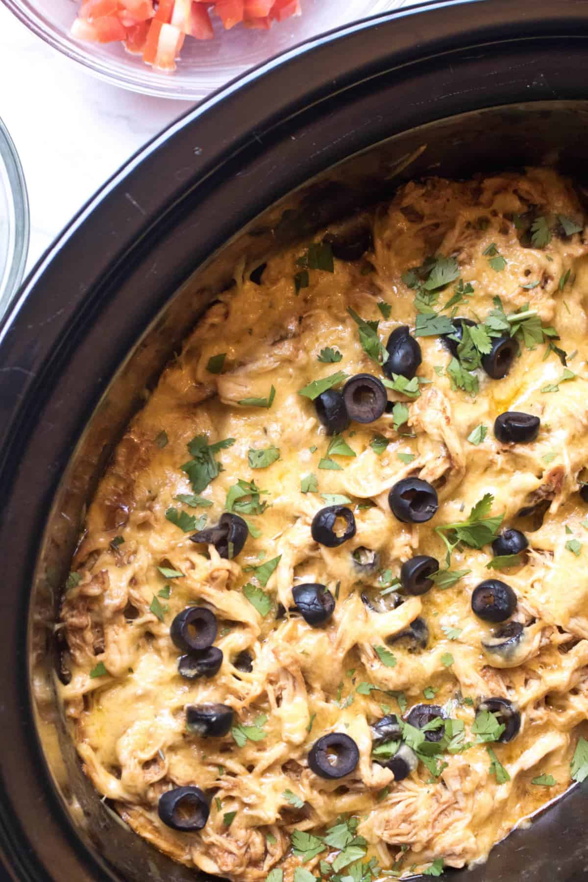 a crock pot with shredded chicken, sliced olives, fresh cilantro, and melted cheese.