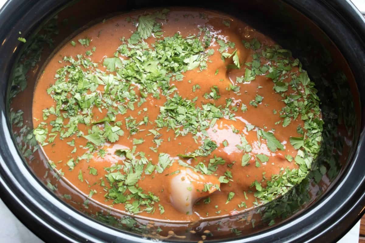 a crock pot with raw chicken covered with enchilada sauce and fresh cilantro.