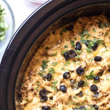 crock with shredded chicken enchilada topped with cheese and sliced olives