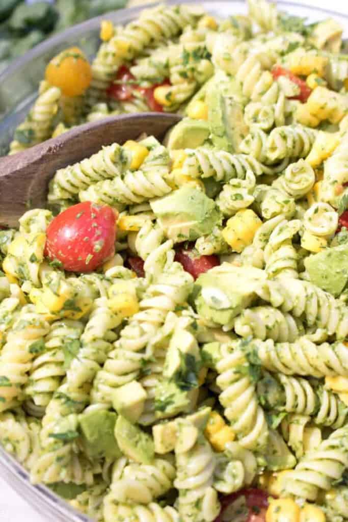 green pasta salad with tomatoes and corn with a wooden spoon in it.