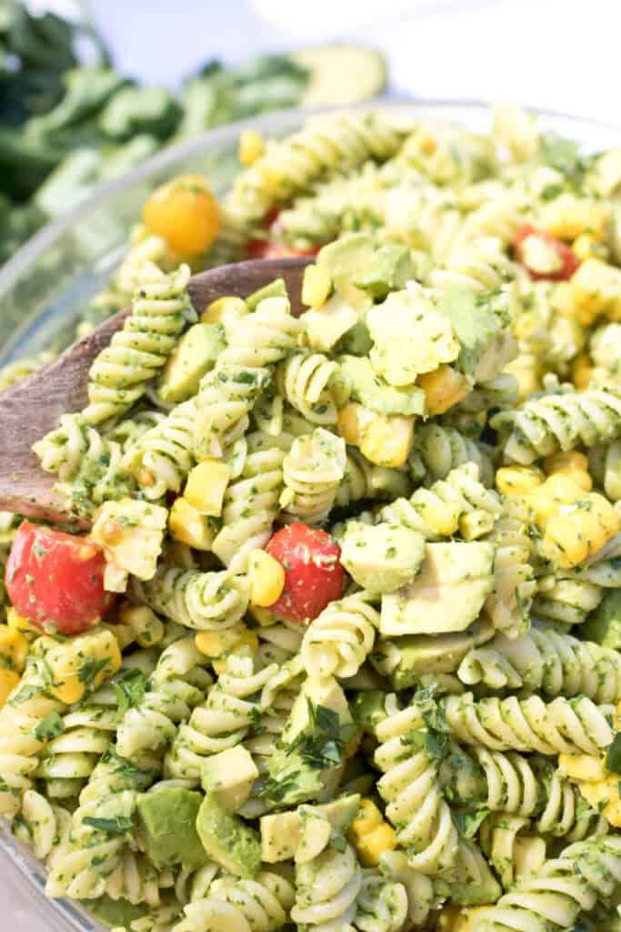 green pasta salad with tomatoes and corn with a wooden spoon in it.