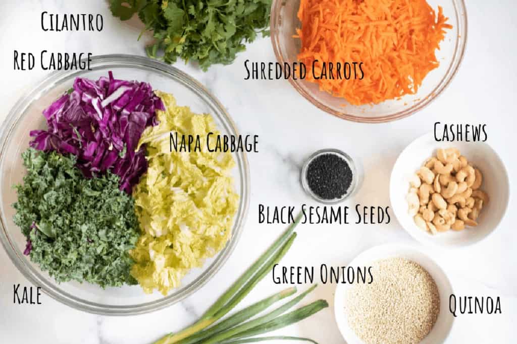 a bowl of cabbage, carrots, onions, kale, cashews, sesame seeds, on a counter.