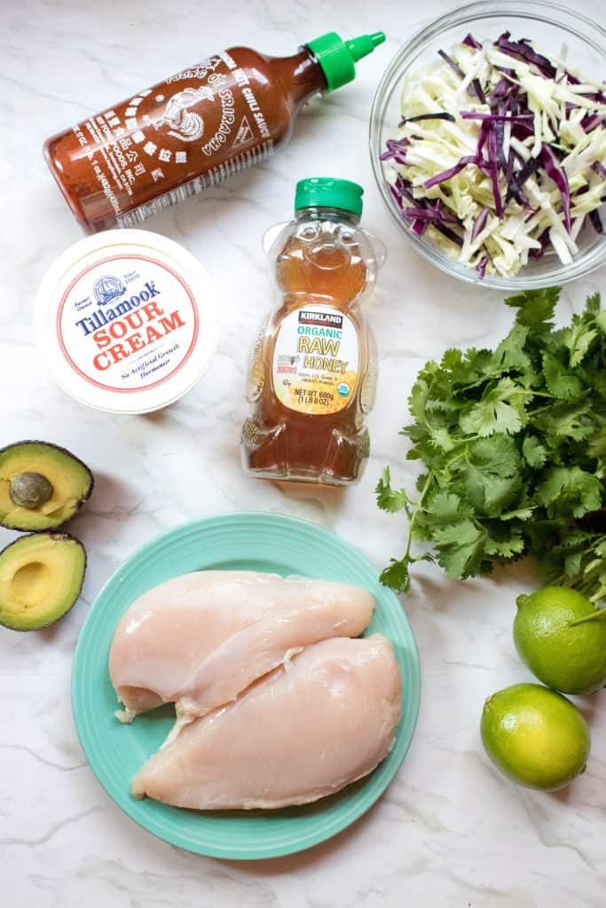 sriracha, cabbage, raw chicken breasts, honey, avocado, sour cream, lime, cilantro on counter.